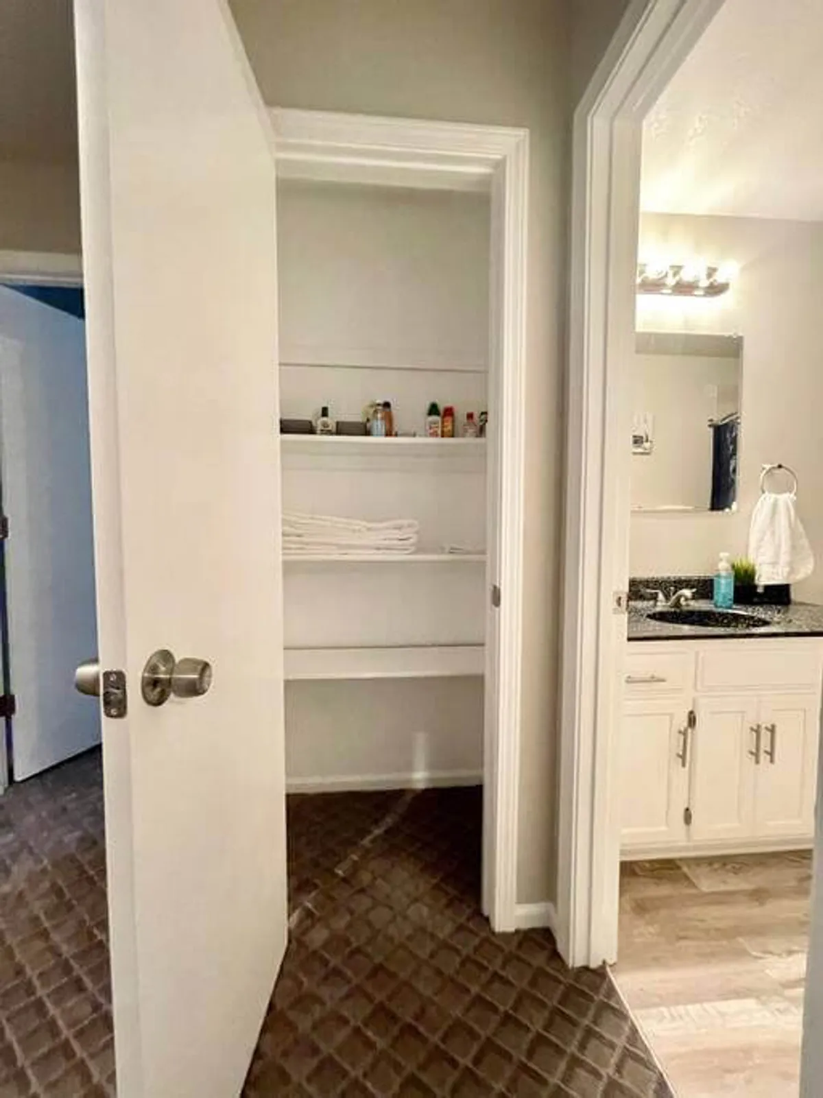 The image shows an open closet door leading to shelves lined with towels and toiletries, plus a glimpse of a vanity area in the bathroom. The space feels tidy and well-supplied, with essentials like fresh linens and cleaning products on display.