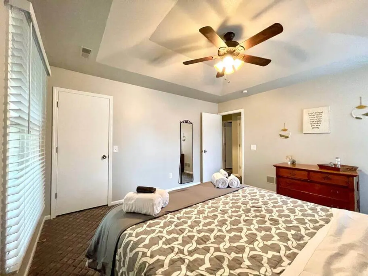 This image features a serene bedroom with a bed adorned with patterned bedding, a decorative mirror, a wooden dresser, wall art, and a ceiling fan. Soft natural light filters into the room, creating a cozy, inviting atmosphere ideal for unwinding and restful nights.