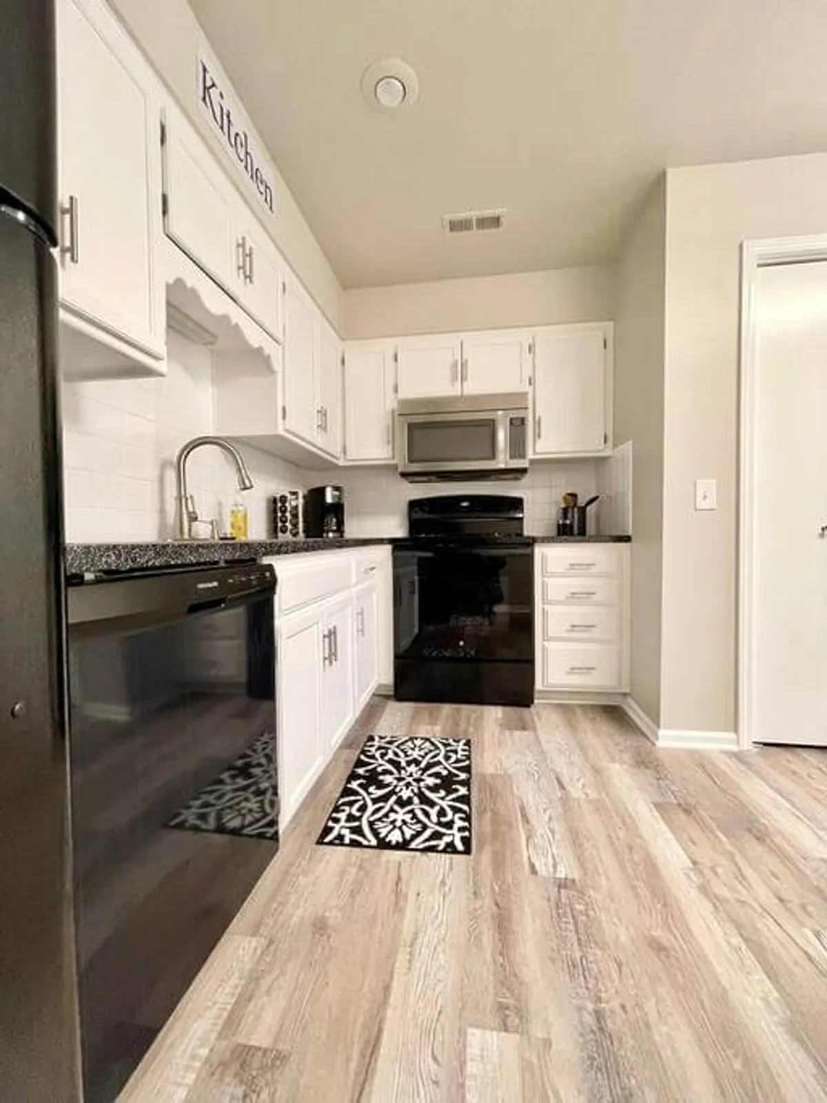 A cozy kitchen featuring black appliances, white cabinets, granite countertops, and a decorative mat. The space is clearly designed for cooking, baking, and hosting family gatherings, with all the essentials for preparing both everyday and special meals.