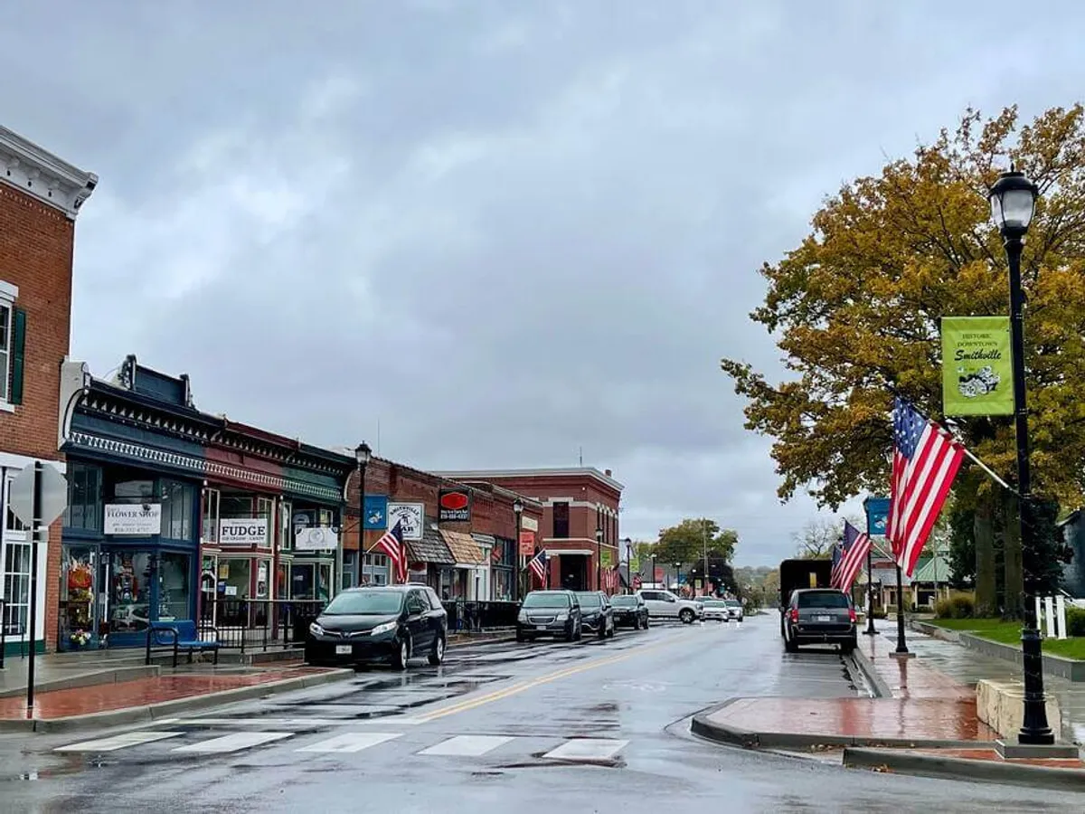 This exterior image captures a charming downtown street in Smithville, featuring historic brick buildings draped with American flags, a tree with vibrant autumn leaves, and locals strolling along the sidewalks. The inviting storefronts hint at a variety of boutiques, cafés, and shops lining the street.