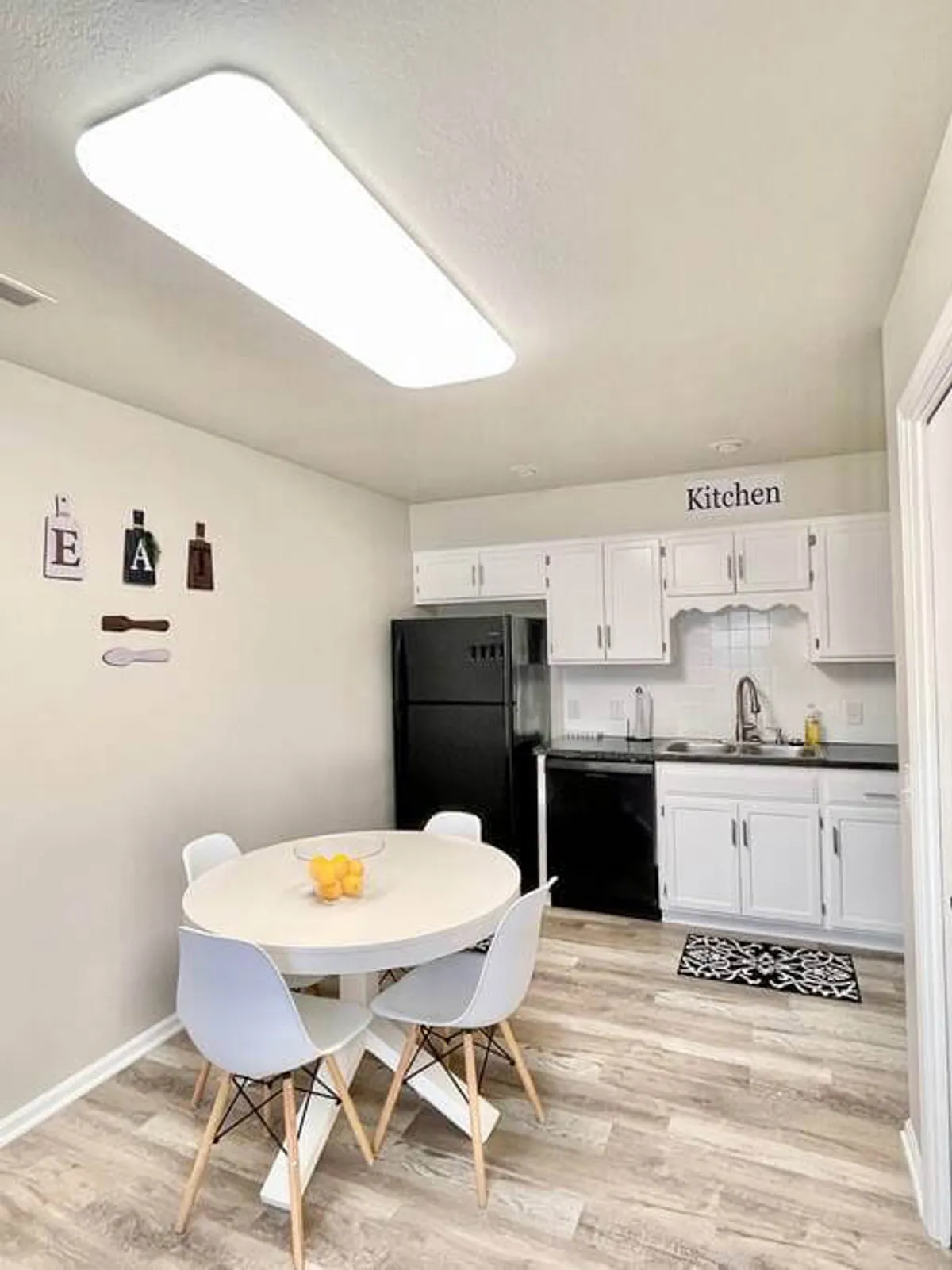 A cheerful dining area with a round table adorned with bright yellow lemons, inviting wall decor, and a view of the kitchen’s black refrigerator and white cabinetry. The setting is perfect for sharing meals or enjoying casual gatherings with family and friends.