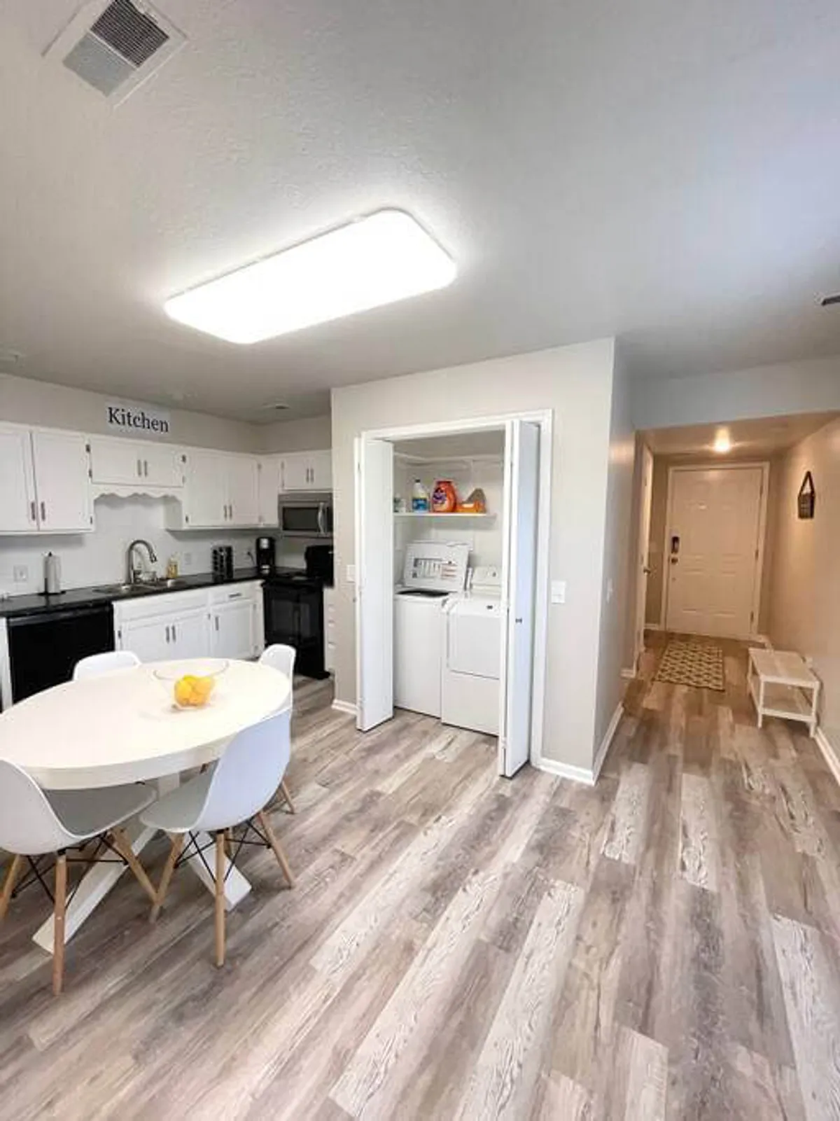 A homey dining and kitchen space featuring a dining table with a fruit bowl, visible kitchen appliances, and a washer and dryer in the background. The area supports multiple family activities, from meals and casual gatherings to multitasking with household chores.