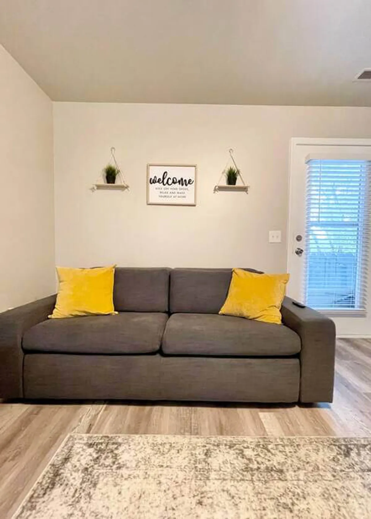 A bright, modern lounge with a gray couch adorned with yellow pillows, a welcoming sign, and shelves decorated with potted plants. The setting suggests a first impression of hospitality and a space primed for casual conversation or solo downtime.