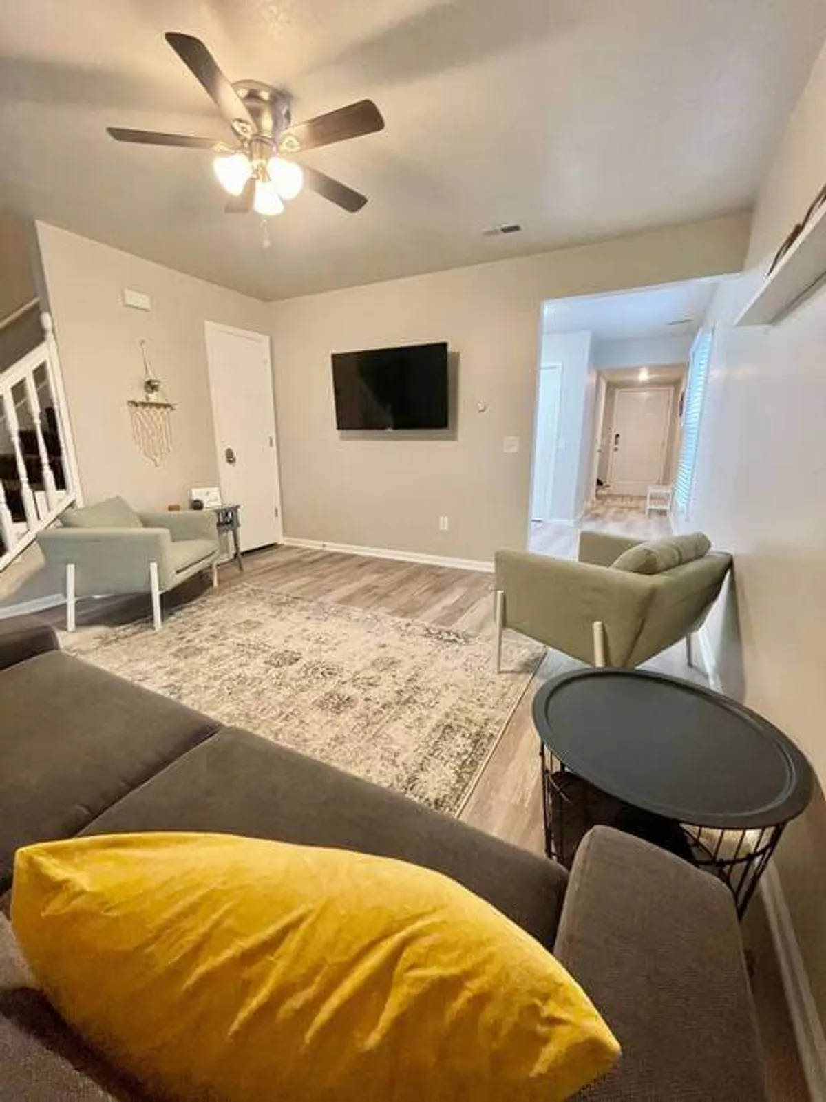 This image centers on a living area with a comfortable sofa accented by a yellow pillow, a large TV on the wall, an area rug, and green chairs. The room is bathed in natural light, creating a welcoming environment ideal for both leisure and entertainment.