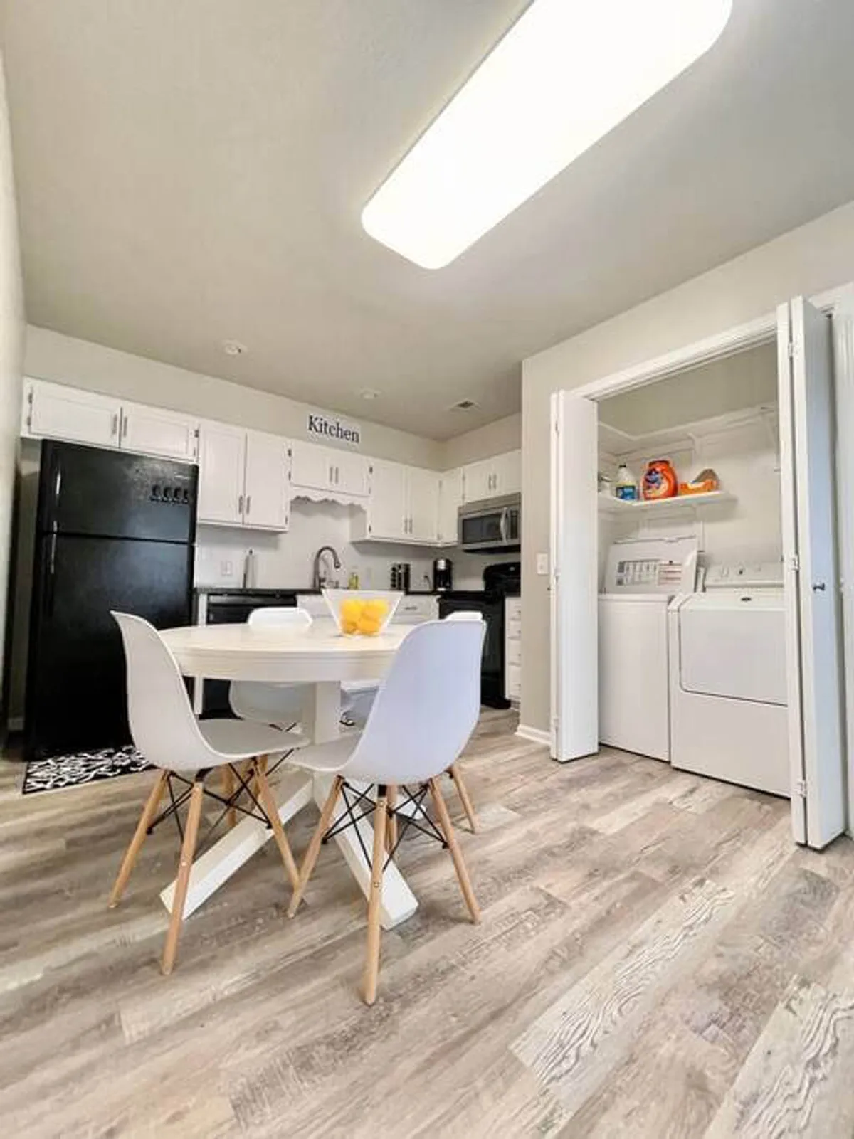 This image features a round dining table with white chairs, visible kitchen appliances, and, crucially, a laundry area integrated into the living space. There's a relaxed, everyday atmosphere, with the laundry area showing clear evidence of a washer and dryer setup.