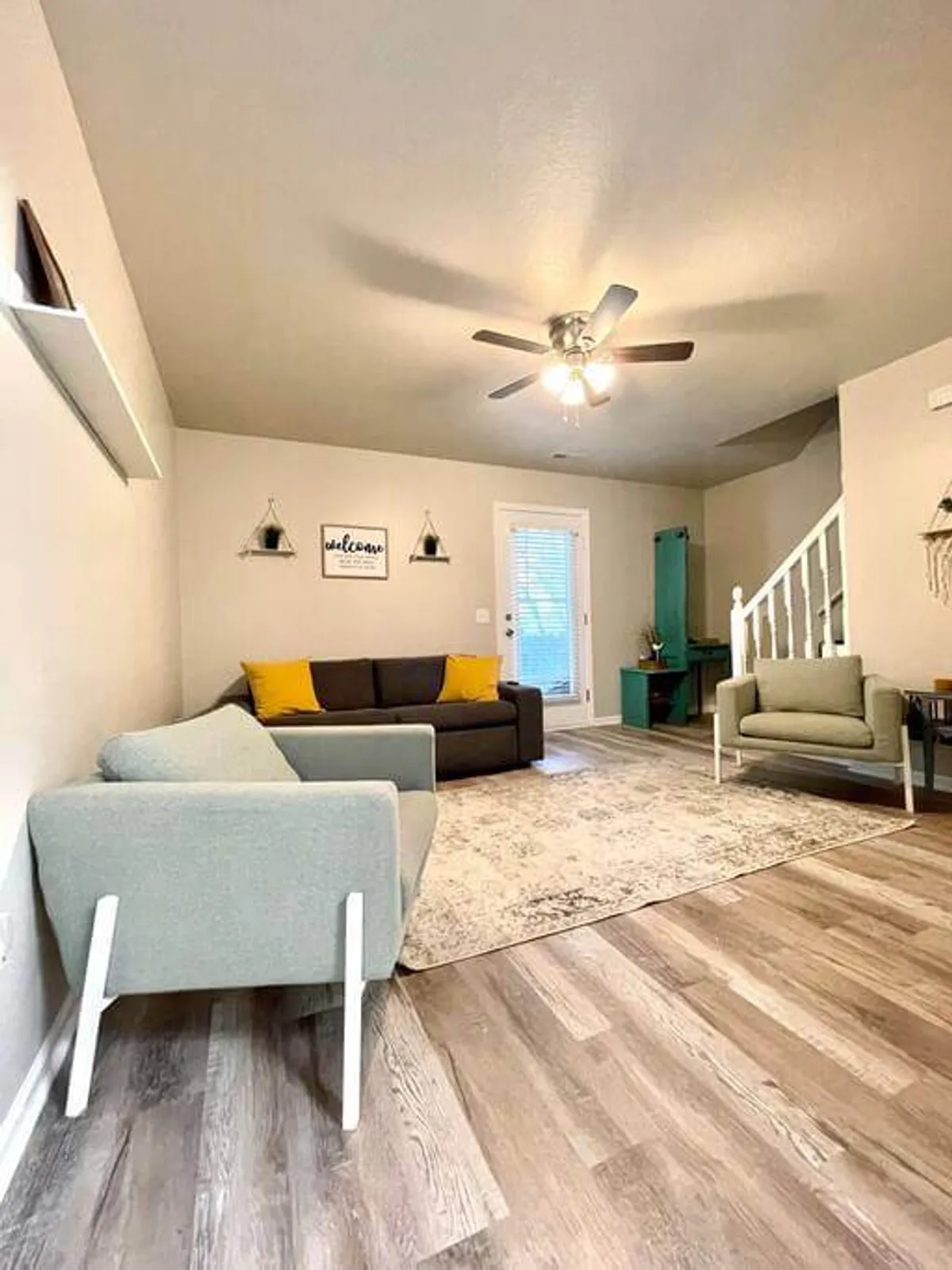 A stylish living area featuring a comfortable sofa, a welcome sign, a decorative rug, vibrant plants, and a staircase leading upward. The space is open and inviting, ideal for both relaxing after work or meeting visitors in a casual yet sophisticated setting.